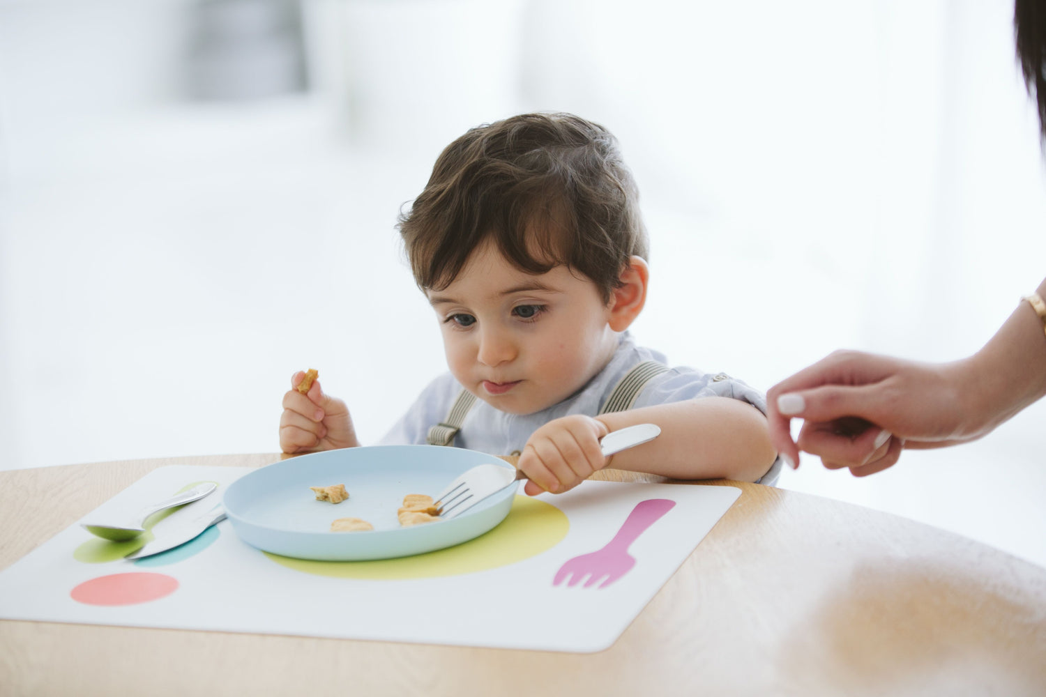 Toddler learning to eat independently with child-sized utensils and a Montessori dining setup.